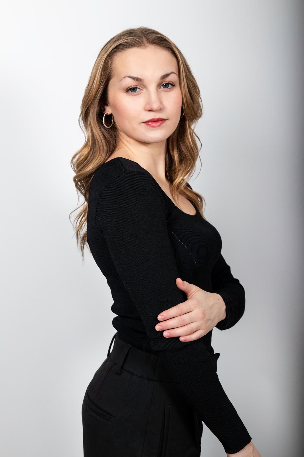 Paige Robinson in black on a pale grey backdrop, looking over her shoulder at the camera
